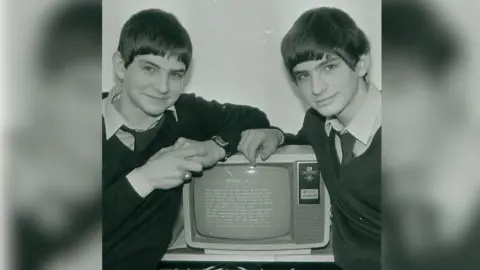 Handout A black and white archive photograph of the Oliver twins as young men, leading on an old computer. They are wearing white collared t-shirts and ties underneath dark V-neck jumpers, smiling at the camera.