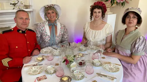 Three women and a man sitting at a table inside Parade House. The table is laid out with afternoon tea, using delicate flowery teacups and champagne glasses. The women are wearing dresses with puffy sleeves and hats, with their hair curled and slightly pulled out at the front. The man is wearing a red army parade tunic with gold detailing.