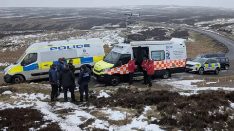 TWSMRT stood with a police van a mountain rescue van. In a separate group, the mountain rescue team can be seen in front of the van in front of the van in a red jacket with black police officers. Morland can be seen behind the van, which is spread beyond the horizon.