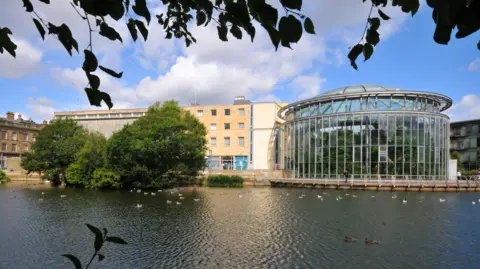 Brick building with multiple windows and curved glass building. There are trees in front of it and a large pond.