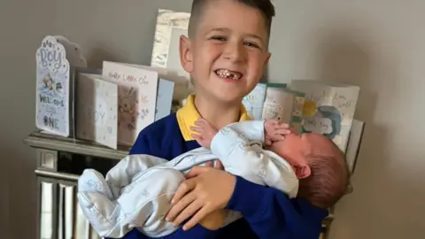 Archie smiles at the camera while holding his baby brother Finley in his arms. Archie has short dark hair and his big grin reveals he is missing his front teeth. He is wearing a school uniform of a yellow polo shirt and blue jumper. Finley has dark hair and is wearing a blue onesie. Behind them is a shelf covered in cards celebrating Finley's birth.