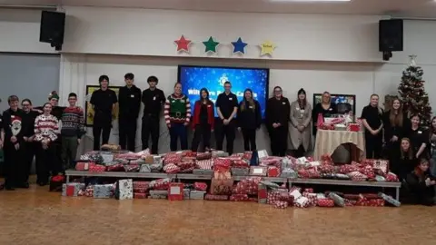 Wiltshire Police A large group of people stand together in a line in what looks like a school hall. There is a large collection of wrapped presents in front of them on a low table. One of the adults standing is wearing an elf outfit and there are four coloured stars suspended on a wall above people's heads. There is also a decorated Christmas tree to the right of the image.