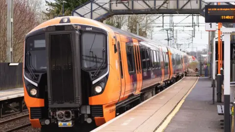 An orange and black Class 730 electric train stopped at a small railway station with a walkway over the track in the background. The platform id deserted. Trees line the other side of the track.