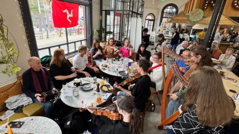 A large group of musicians sat around several tables in a large crowded cafe. There are people playing harps on the right, and many violins, as well as an accordian and flute among the instruments. There is a window on the left with the red Manx flag hanging in it.