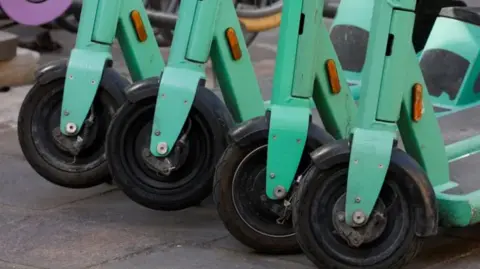 Reuters Four black front wheels of mint green electric-scooters. The wheels are black and the frame of the scooters is mint green. They are lined up in a row on a pavement. 