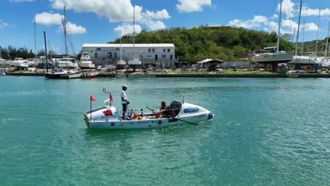 ATLANTIC DASH Mike is sat down rowing, and Tim is stood up on the boat, in turqoise waters as they reach land.