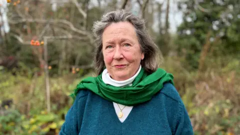 Judith Heinemann with long brown hair, looking at the camera and wearing a blue V-neck jumper over a white top. There is a green scarf around her neck. There are trees and a hedge behind her (in her own garden).