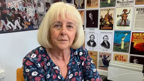 Ruth Jones seated in a wooden chair. She has light-coloured, shoulder-length hair with a straight bob hairstyle and is wearing a blue floral-patterned top featuring pink, white, and light-blue flowers. A small cross pendant hangs from a thin necklace around her neck. The wall behind her is covered with a display of posters, historical prints, and photographs.