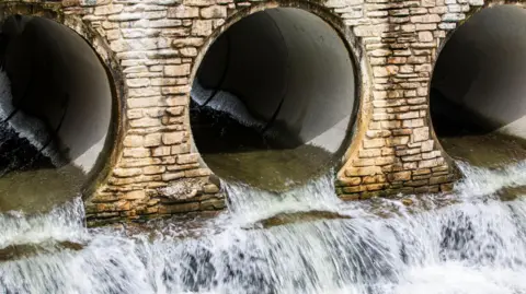 Water flows out of three openings of underground pipes. The pipes are surrounded by lightly coloured brickwork.
