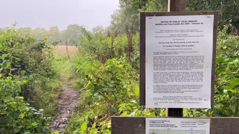 Local Democracy Reporting Service A planning inquiry notice on a post by a footpath. You can see foliage and trees in the distance.