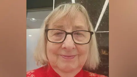 Lorraine Dunn Lorraine Dunn, with short blonde hair and black-rimmed glasses, wearing a red jumper, looks at the camera. 