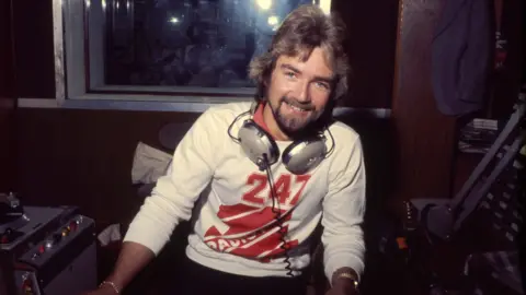 A colour mid-shot of a man sitting, pictured from the waist up, looking at the camera smiling, leaning slightly towards the right-of-frame. He has long light brown hair and a similar-colour beard, and is wearing a white jumper with a large red Radio 1 logo on it. He is sitting in a 1970s radio studio with various analogue equipment visible, and has a pair of heaphones slung around his neck. Behind him is a window through to the control room area of the studio.