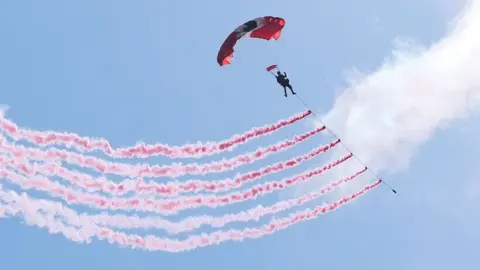 PA Media A single parachutist is high in the sky beneath a large rectangular parachute canopy. The parachute is mostly red with dark grey and white sections near the centre. The person appears as a dark silhouette suspended by thin lines connecting to the canopy. Trailing from beneath the parachutist is a long line supporting several smoke emitters. These emitters create five parallel streams of pinkish-red smoke that stretch diagonally across the image from upper right to lower left. The smoke trails are wavy and textured, resembling broad brushstrokes painted across the sky. The background is a bright, clear blue sky with a large patch of white cloud on the right side.