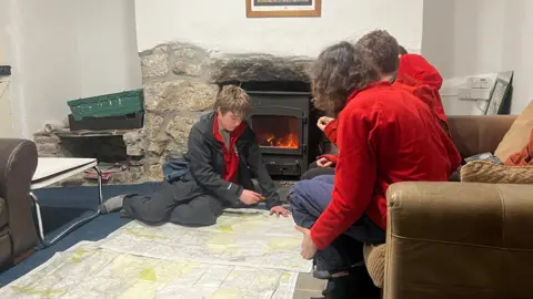 Students looking at large maps on the floor in front of a fire. Two people in red fleeces are sat on a sofa, while Max is sat on the map with a pen planning their routes. 