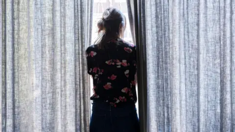 Getty Images A woman is looking through closed curtains through the window. She has dark hair tied back in a ponytail and a navy shirt with a floral pattern on it. This is a creative image and not one of Sophie.