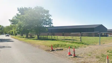 A Streetview image of a road with a large industrial building to the right. A track off the road is blocked by wire link fence and a few orange cones.