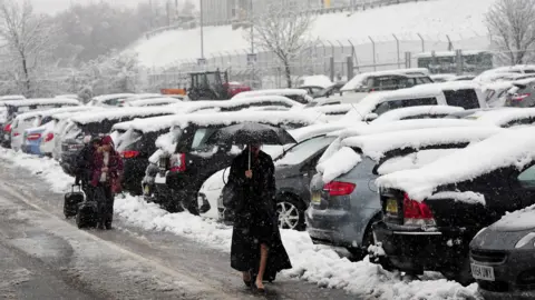 Woman walking through a car park in heavy snow carrying an umbrella