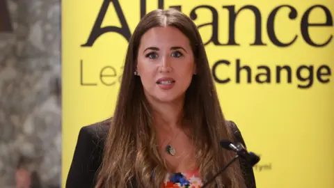 PA Media Sorcha speaking into a microphone. The background behind her is yellow. She is wearing a black blazer and multi-coloured top. She has long straight brown hair.