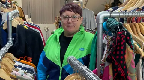 KATHERINE MAYER Woman with dark purple hair wearing a fluorescent green and blue jacket among rails of clothes