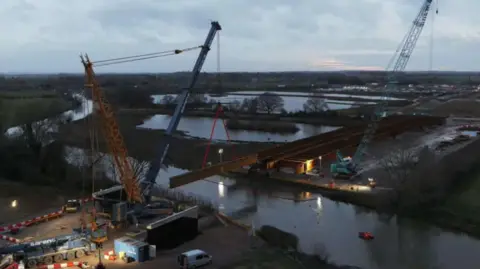 National Highways Cranes lift beams into place across a river. The work is happening under floodlit conditions