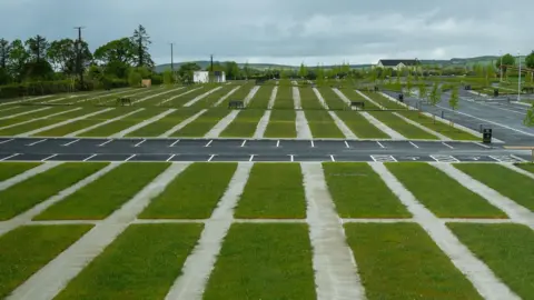 DCSDC A wide shot of the new cemetery. A row of grassy areas are visible with a number of parking spaces in the background. 