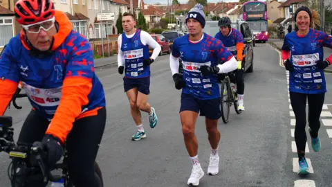 Kevin Sinfield and his support team run down a road in Filton, Bristol, as part of his 7 in 7 charity challenge from Gloucester to Bristol. The runners are wearing blue tops and behind them is a car and then a bus