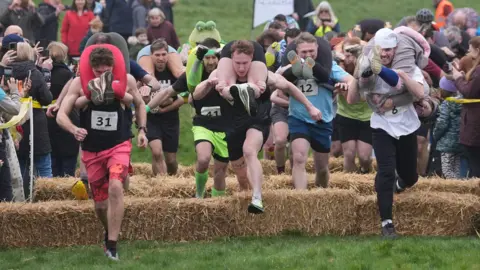Several men race with women hanging from their backs. 