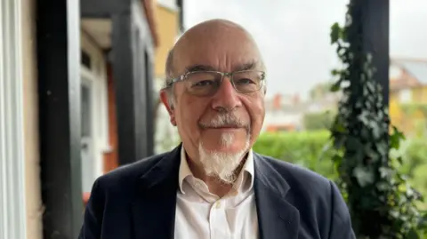Anthony Sheppard, the chair of Claygate Parish Council's planning committee, looks straight at the camera. He is wearing a white shirt and a dark suit jacket, and has a small white beard.