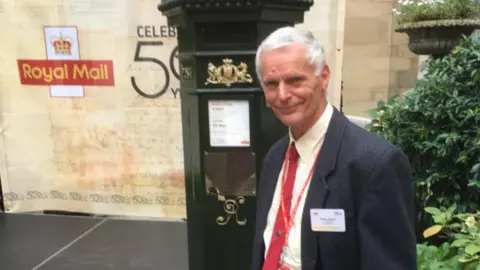 PA Media Postman Robert Hudson stands in front of a special postbox to mark 50 years of the royal mail. Robert is wearing a suit with a red tie.