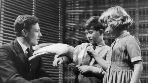 Getty Images A black and white image of two children and man in a suit. The boy holds a white cockatoo on his arm whilst all watch on. 