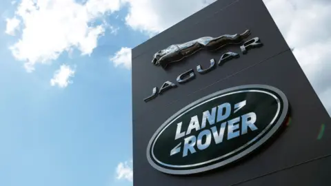 The image is pointing upwards with a tall, dark-coloured sign with the silver Jaguar logo above the green Land Rover logo in the foreground. Above the sign is blue sky dotted with white clouds.