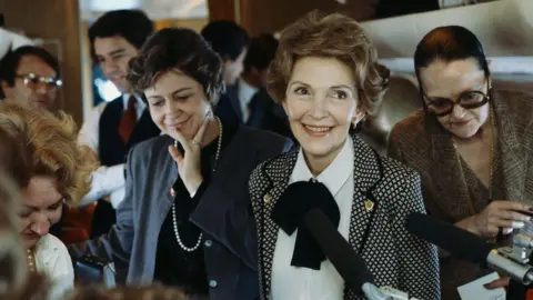 Getty Images/Wally McNamee Then-First Lady Nancy Reagan is smiling, wearing a polka-dot blazer. She stands in front of busy people in the background, looking at the microphones being held towards her.