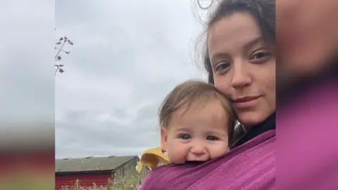 Mother Hannah Howell (right) has brown hair and blue eyes and looks into the camera. While her 7-month-old baby with blue eyes and brown hair strapped to her chest smiles into the camera. 