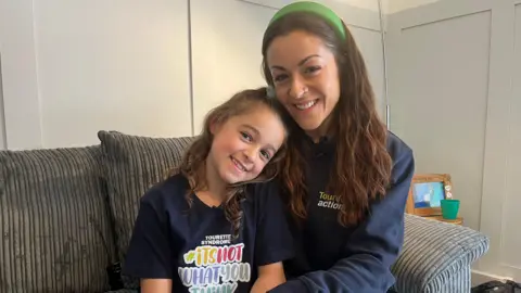 BBC Freya and her mum Louise sit on a grey sofa. Freya is wearing a navy blue t-shirt which has the Tourettes Action logo. Louise is wearing a navy blue hoodie with the Tourettes Action logo. They both have brown hair and Louise is wearing a green hair band. 