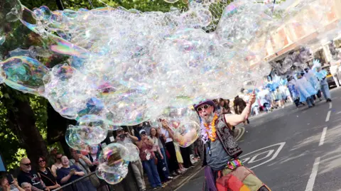 Paul Norris A bearded man wearing colourful patchwork trousers, round sunglasses, a purple hat, leather studded waistcoat and floral garlands, creates a huge bubble through a net, delighting the crowd watching at the side of the road.