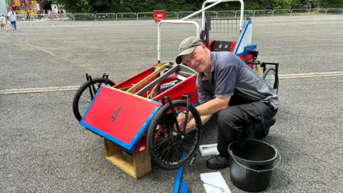 Mark Grice knelt down next to a blue and red homemade kart. He is wearing a grey cap, blue and grey polo T-shirt, black trousers and black boots. The kart is parked on a road. 