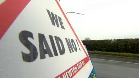 A red and white triangular sign on a rural road says "We said No".
