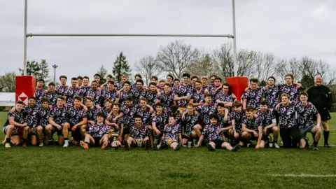 Cotswold Pictures A large group photo of dozens of people who took part in a charity rugby match. They are posing on a rugby pitch in front of posts.