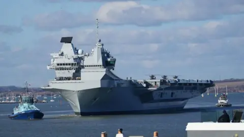 PA Media A large military ship on the ocean with two smaller ships on the water on either side