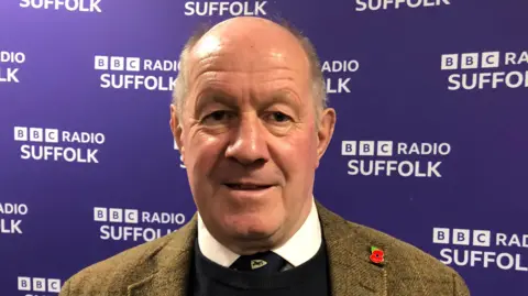 Vikki Irwin/BBC Tim Passmore smiles at the camera while standing in front of a purple coloured wall with the BBC Radio Suffolk logo written on it. He is largely bald with some grey hair and he wears a tweed jacket with a black jumper and white shirt and tie underneath.