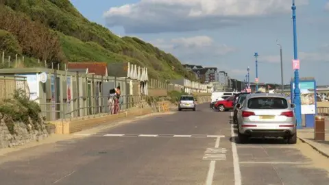Malc McDonald A road bordered bordered by a sandy beach on one side and beach huts and a sloping cliff with vegetation on the other. Cars are parked along the beach side to the right.