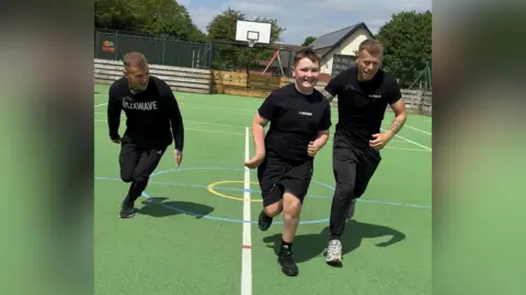 Smiling Liam running across an outdoor basketball court, being encouraged on either side by Harry and Joe Freeman, founders of the Boxwave fitness programme.
