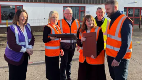 Six people are standing outside a factory. On the left is a woman in a purple high-viz jacket, to her right are two men and two women - including Michelle O'Neill who is blonde and Caoimhe Archibald who has auburn hair - wearing orange high-viz. O'Neill and Archibald are holding up MDF panels. A third man further back is wearing yellow high-viz.