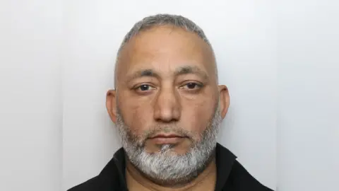 A mugshot of Muhammad Arshad. He has brown eyes and a white and grey beard. He is wearing a black top with a collar. 