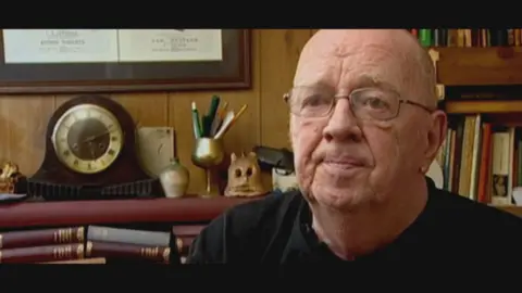 Ian McNicol in 2007 wearing a black tshirt and silver rimmed glasses. In the background there is a bookshelf, piles of books and a clock.