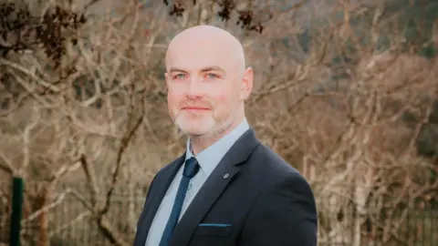 Jody Mussen  A man wearing a dark blue suit and blue tie is standing in front of a fence and some bare trees. He is looking directly at the camera. 