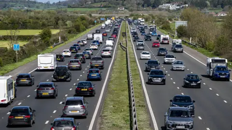 Ben Birchall/PA Wire Image shows rows of traffic on the M5 heading northbound and southbound. There are several cars travelling in both directions including caravans, 4x4s and smaller vehicles. Image appears to be captured from a bridge above the road and shows the central reservation in the middle.