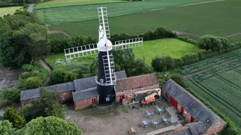 Joe Bilton/BBC An aerial picture of two sails placed horizontally on a windmill. The sails are white and they are placed on a white windmill cap. The windmill is in a courtyard surrounded by farm buildings and green fields.