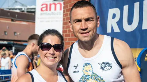 PA Media A tall muscly man, Kevin Sinfield, in a white running vest. He has short hair and is smiling at the camera. On his left is Lindsey Burrow, also smiling. She has brown hair in a ponytail and sunglasses on. Behind them are people milling about before the start of the run.
