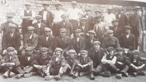 Memories of Eye Facebook page Black and white photo of boys and men lined up in four rows at Dogsthorpe brickworks, Peterborough, in about 1900, all wearing flat caps